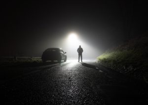 A road at night with a dark car parked on the side and a person wearing a black hooded jacket standing under a light next to the car.